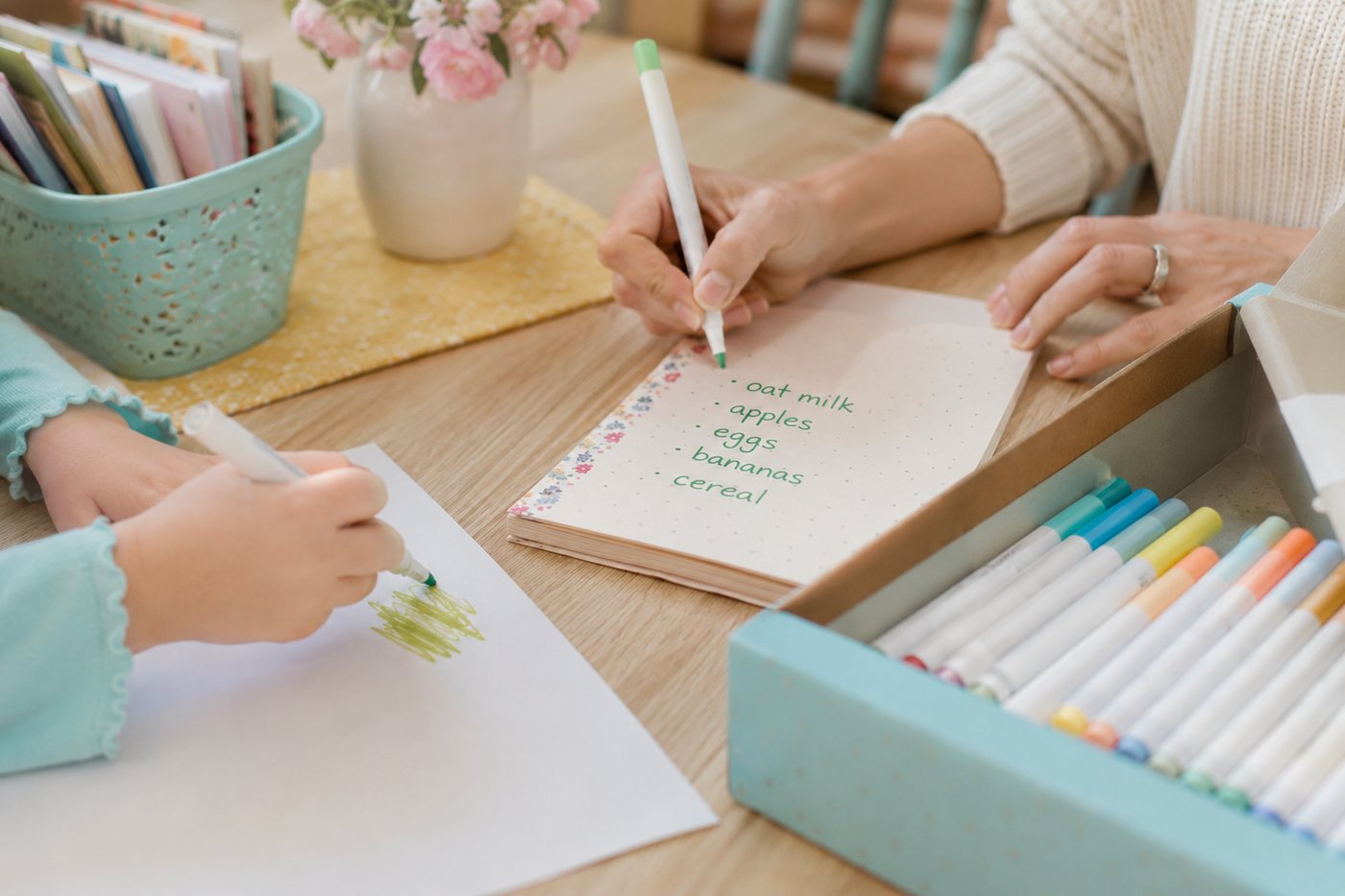 An adult writing a shopping list in a dotted notebook while a child colours.