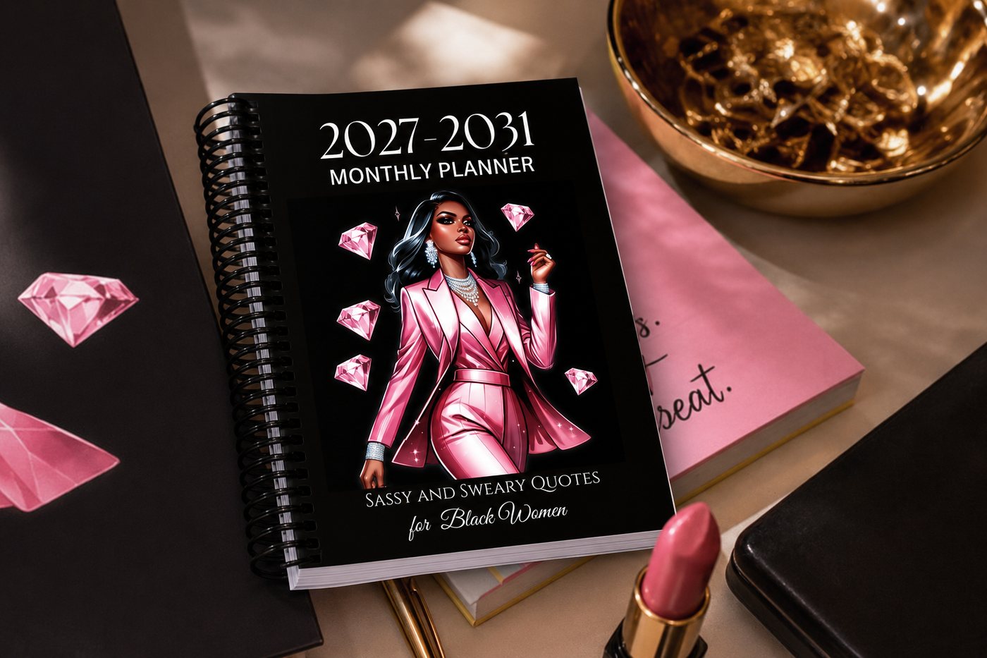 The 2027–2031 Sassy and Sweary Monthly Planner on a desk beside a gold dish and a pink notebook.